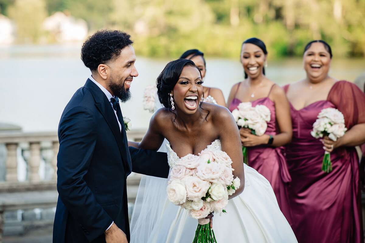 A candid moment of joy as the bride and groom laugh heartily, surrounded by bridesmaids in matching burgundy dresses, with a waterfront backdrop