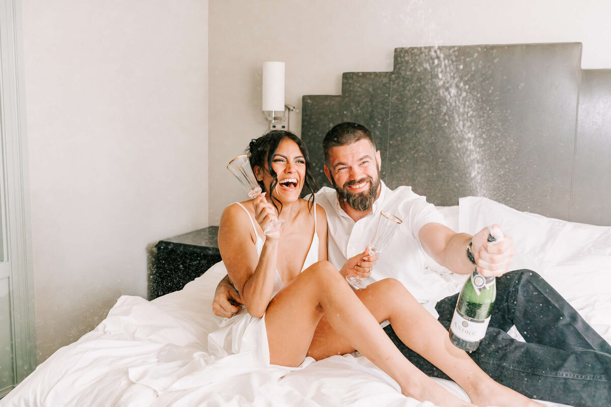 bride and groom pop a champagne bottle in hotel room to celebrate their engagement