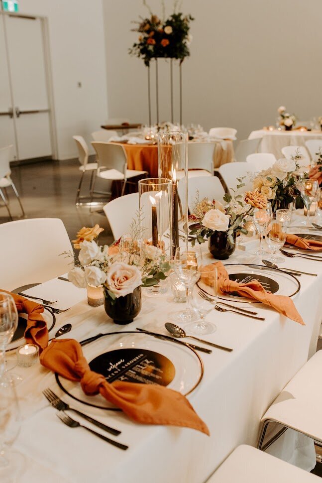 This beautifully arranged reception table at the Ottawa Art Gallery features long white tables adorned with black cutlery and modern black rimmed charger plates. Each setting showcases a sleek black menu and a knotted terracotta napkin, complemented by lit black tapered candles and charming earth-tone floral arrangements, ideal for couples seeking stylish decor for their wedding or event.