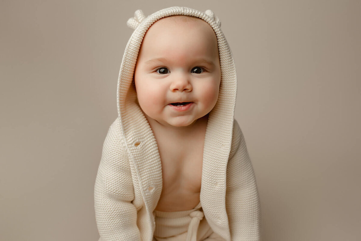 a baby wearing a teddy bear hood smiles at the camera on a white drop