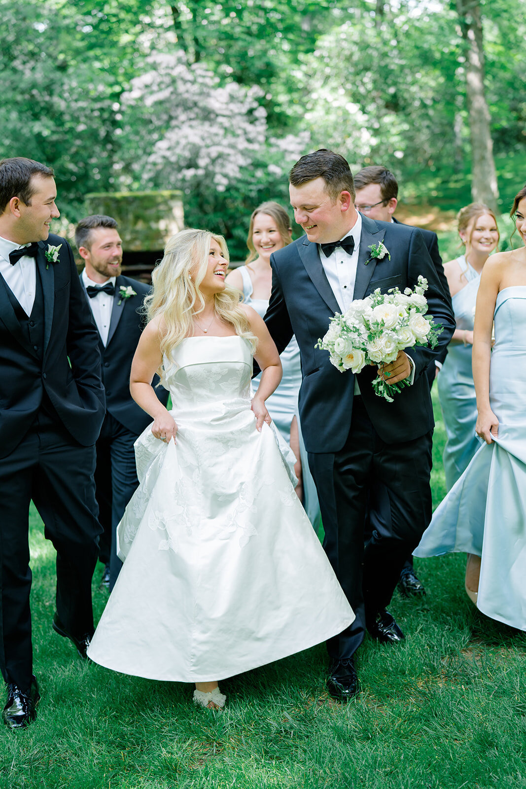 Wedding party walking together outside, bride and groom smiling while surrounded by bridesmaids in light blue dresses and groomsmen in black tuxedos.
