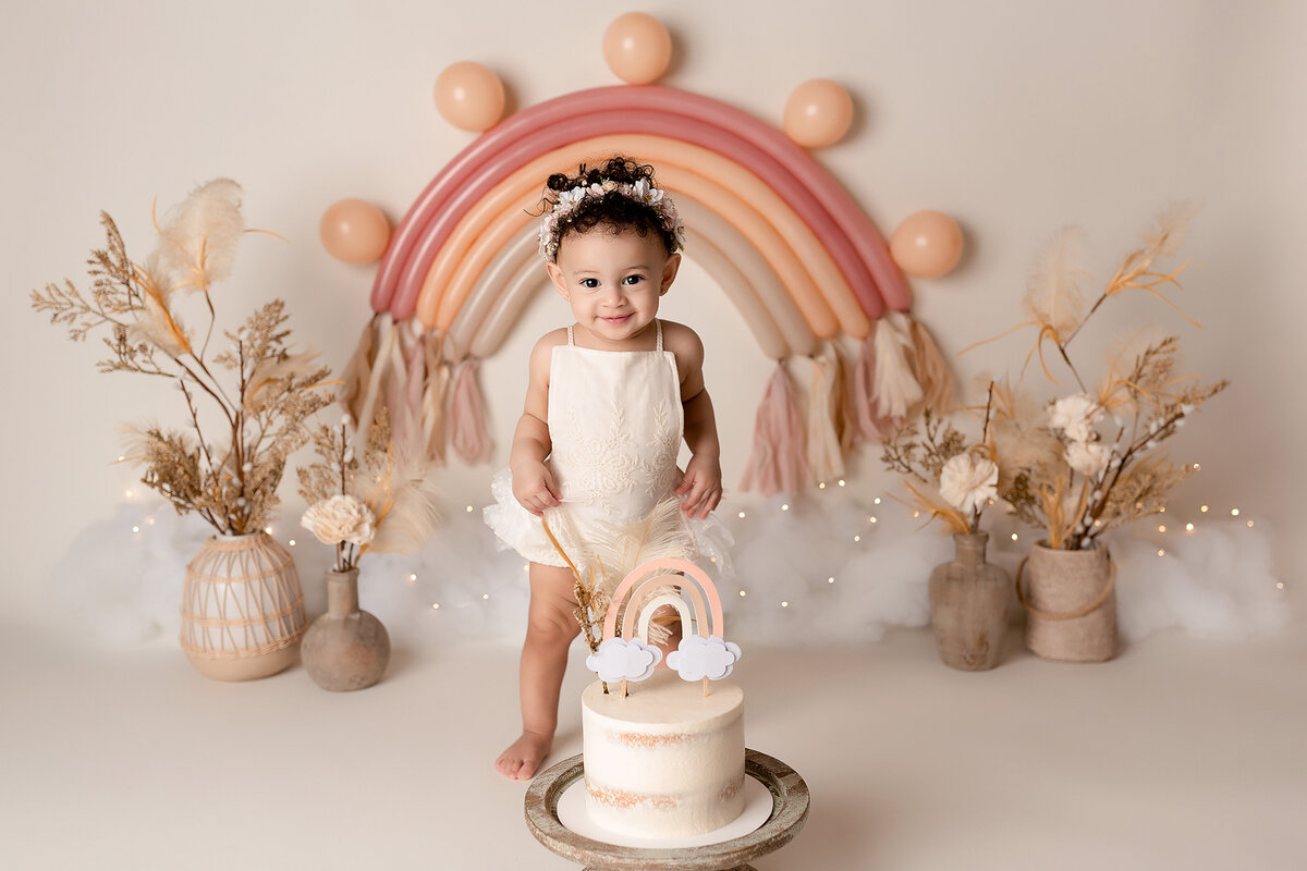 Vintage boho themed cake smash with top West Palm Beach and Jupiter, FL photographer. Baby is standing behind cream cake with a rainbow smiling at the camera wearing a white romper. Behind her, there is a monochromatic pink, peach, and tan rainbow with faux clouds. There are grasses, flowers, and feathers in shades of cream and taupe.
