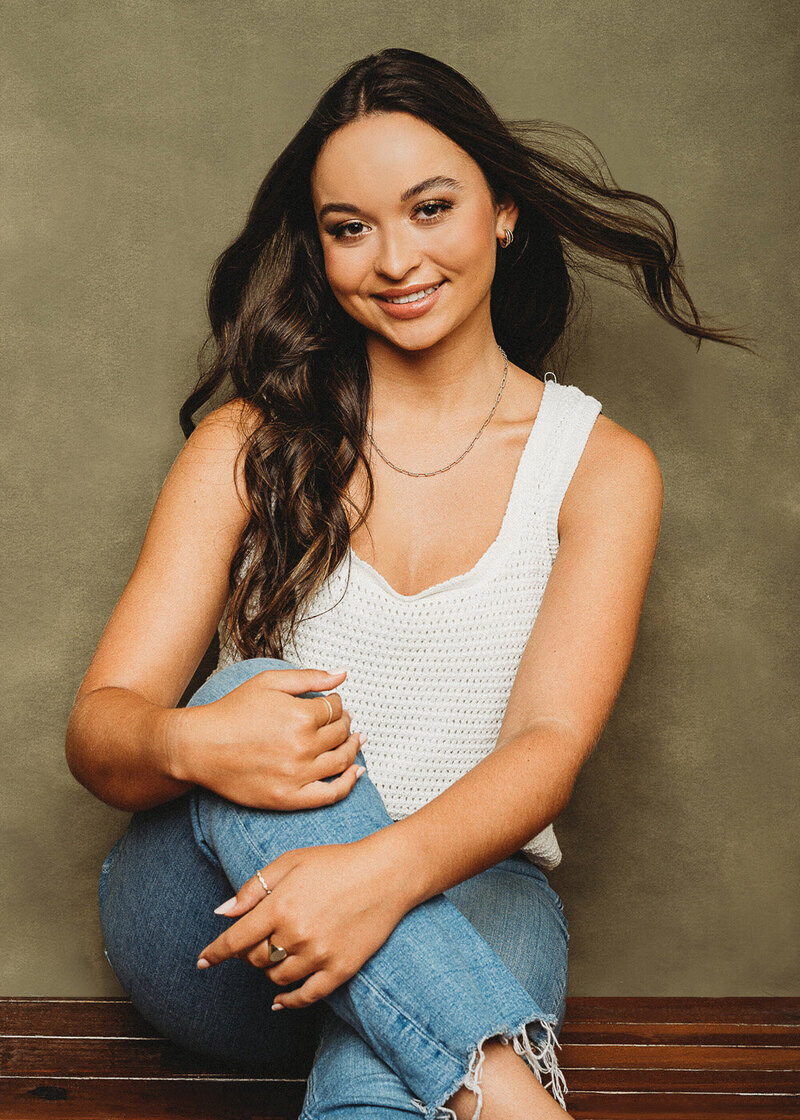 Here is another example of a high school senior photoshoot Charleston SC. This photo depicts a a young women in jeans and a white tank top smiling as she is seating in the photo studio. With a fan blowing her dark brown hair slightly for an added touch of elegance.