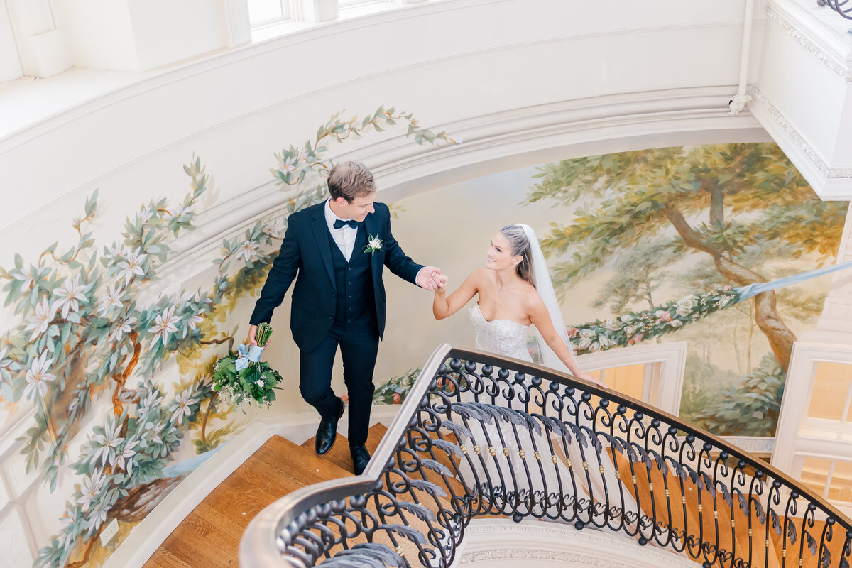 Gentle groom helping his beautiful bride up stairs inside a mansion with a stunning floral mural behind them at The Olana Wedding Venue