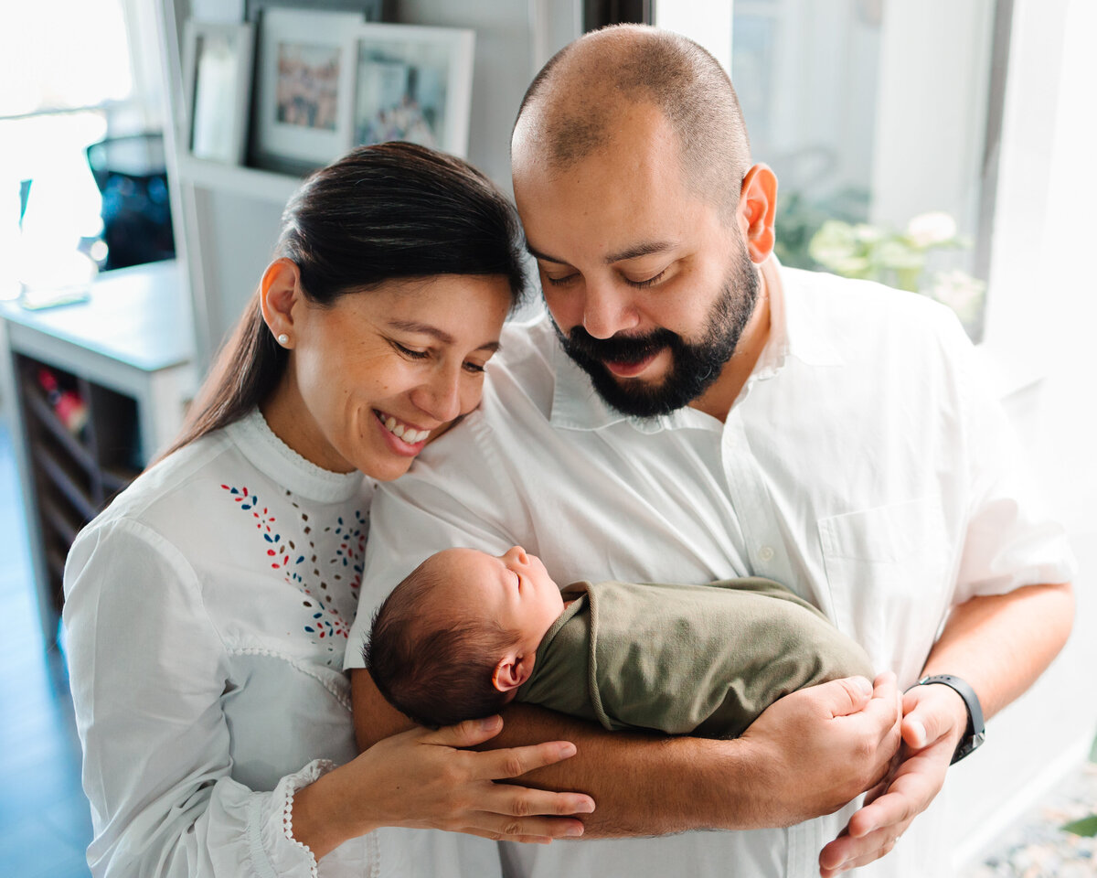 Maternity photography packages in Albuquerque with this heartwarming family photo. The parents, dressed in white t-shirts, gaze lovingly at their newborn baby, who is wrapped in a cozy green blanket. The background features portraits and a fireplace, adding a homely and intimate touch to the scene.
