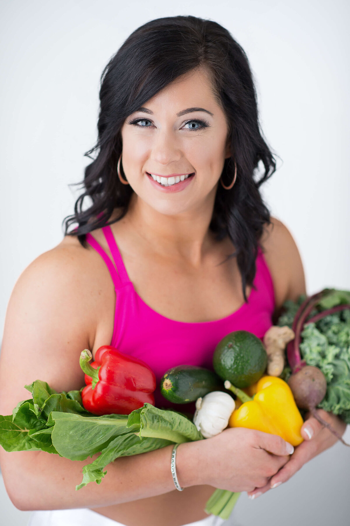 a female personal trainer in a pink tank top holding a bouquet of assorted vegetables.  Captured in studio by Ottawa Branding Photographer JEMMAN Photography COMMERCIAL