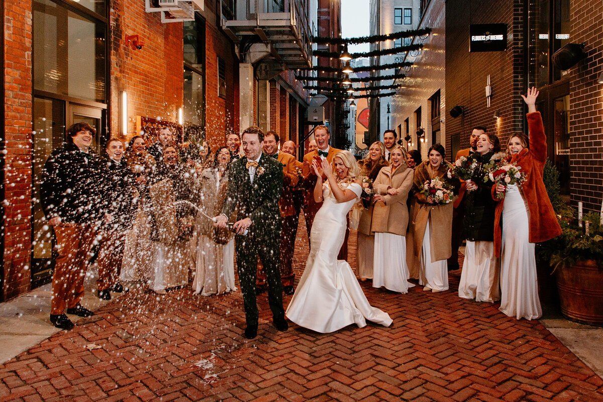 a wedding party cheering as the groom and brdie pop and spray a bottle of champagne  in the middle of a narrow brick city alley