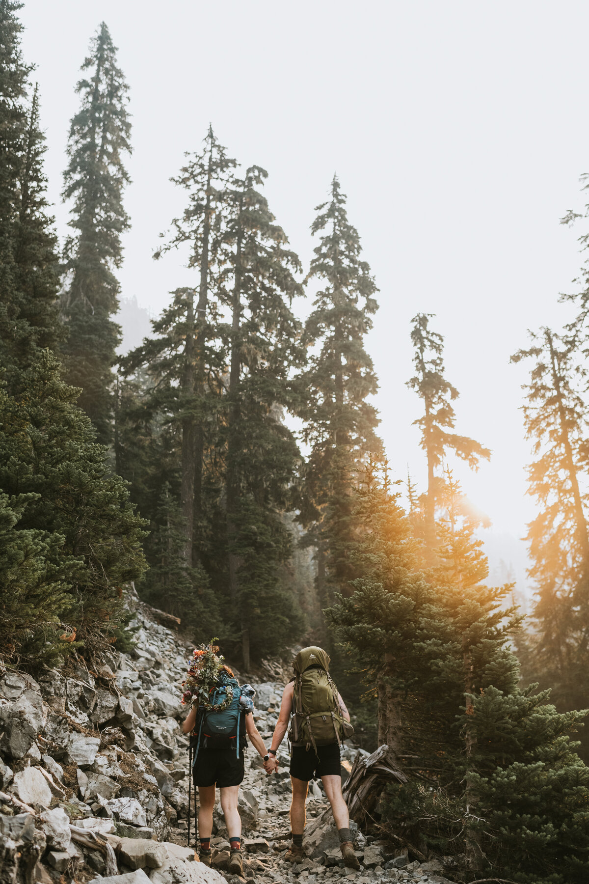 a couple hiking with their wedding attire and flowers in their backpack surrounded by trees as they go to a glacier lake for their elopement day