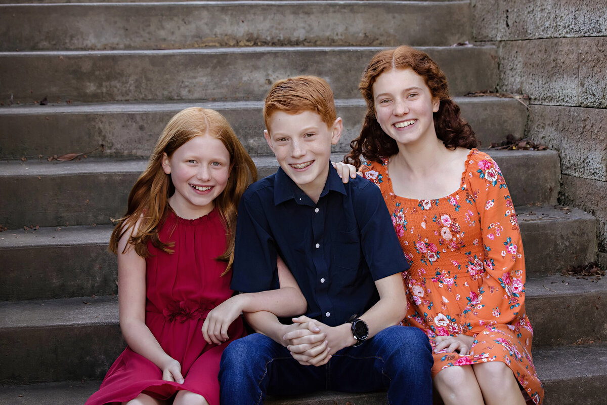 Striking autumn photo of three siblings with red hair seated on weathered concrete staircase at Ballantyne Corporate Park Charlotte NC