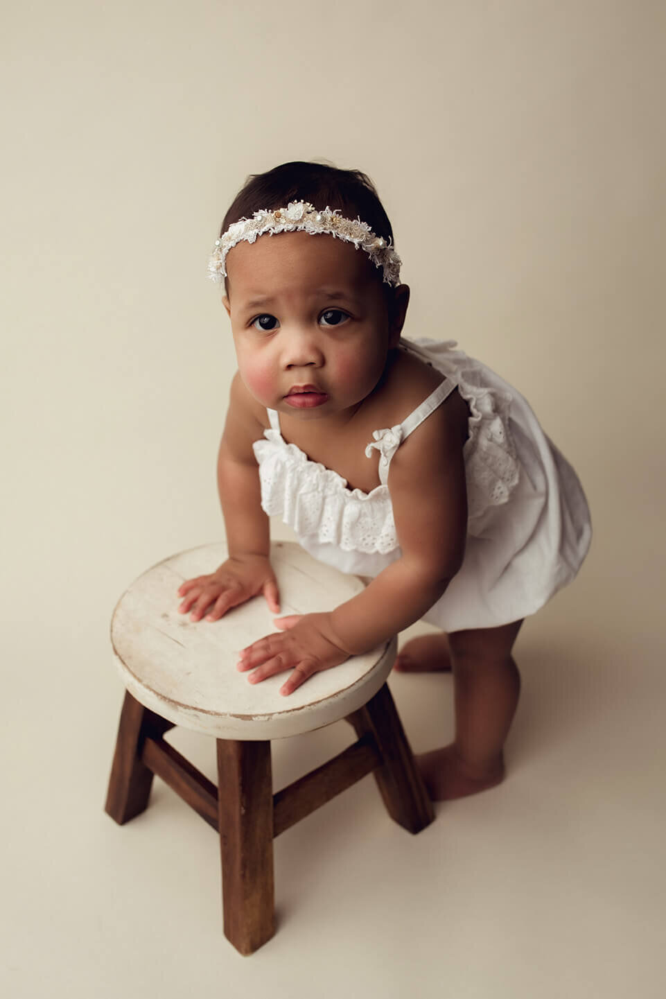 a one year old girl being very serious while holding onto a stool