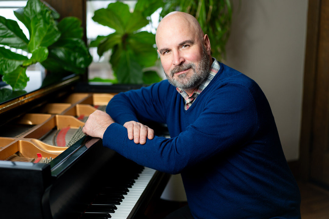 Professional headshot of man in blue sweater seated at piano smiling warmly with plants background new jersey.
