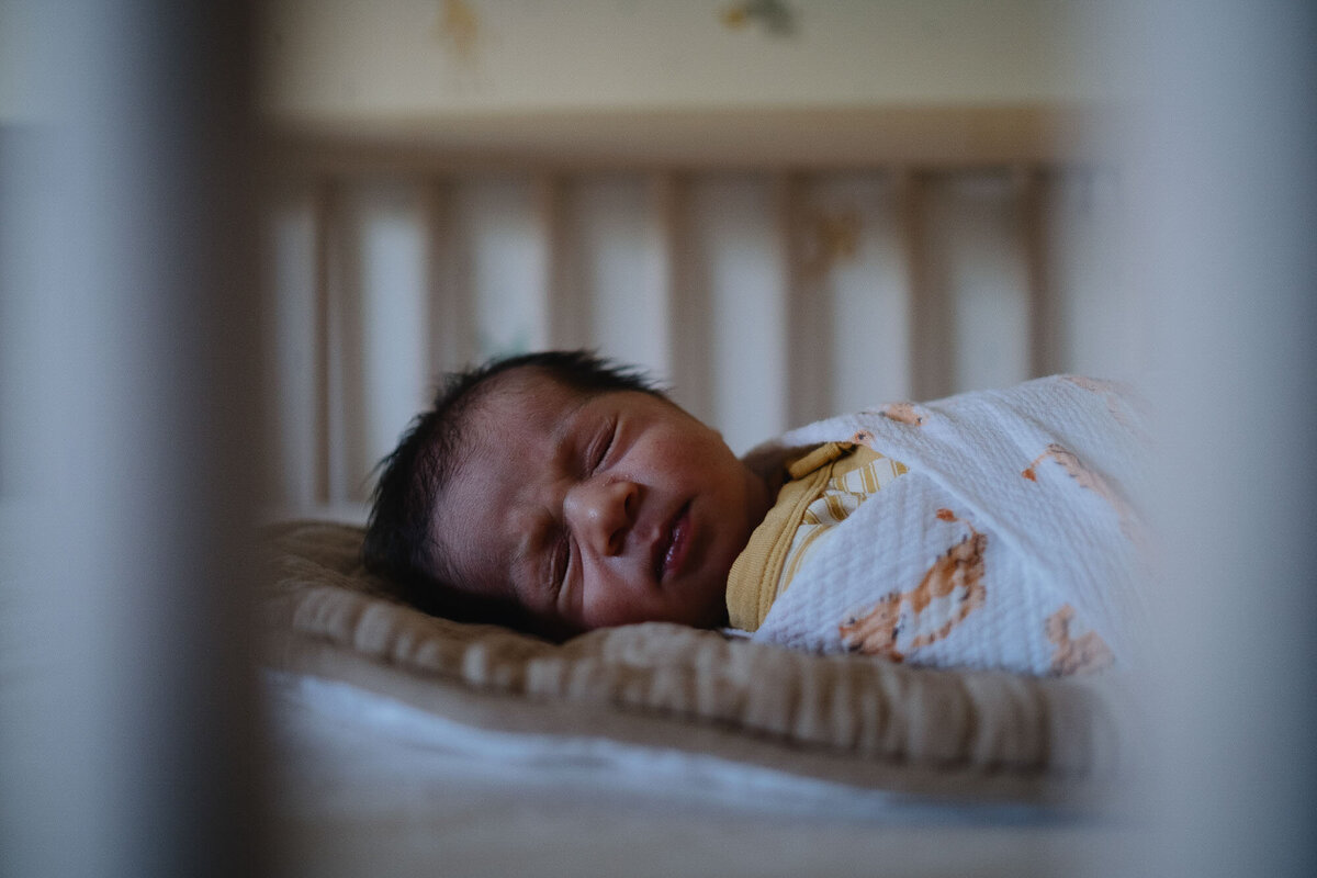 A newborn baby boy sleeps in his crib, swaddled in a white and orange blanket.