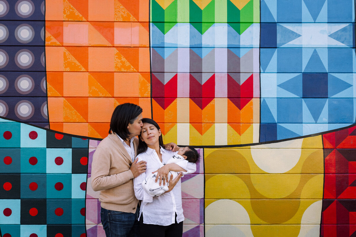 mom in white shirt holding newborn wrapped in white swaddle while dad in tan sweater stands next to and kisses mom on head in front of colorful San Francisco mural