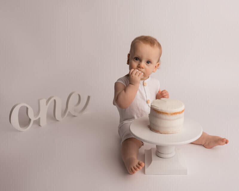 Baby boy in a white romper sitting next to a small cake with the word "one" in the background.