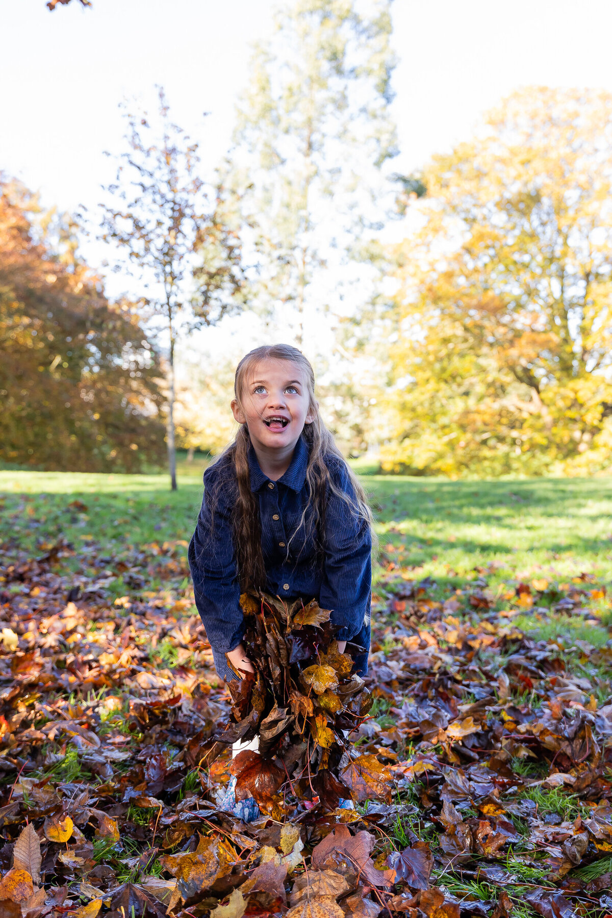 Young girl in a denim dress playing with autumn leaves in a park, looking upwards with a joyful expression.