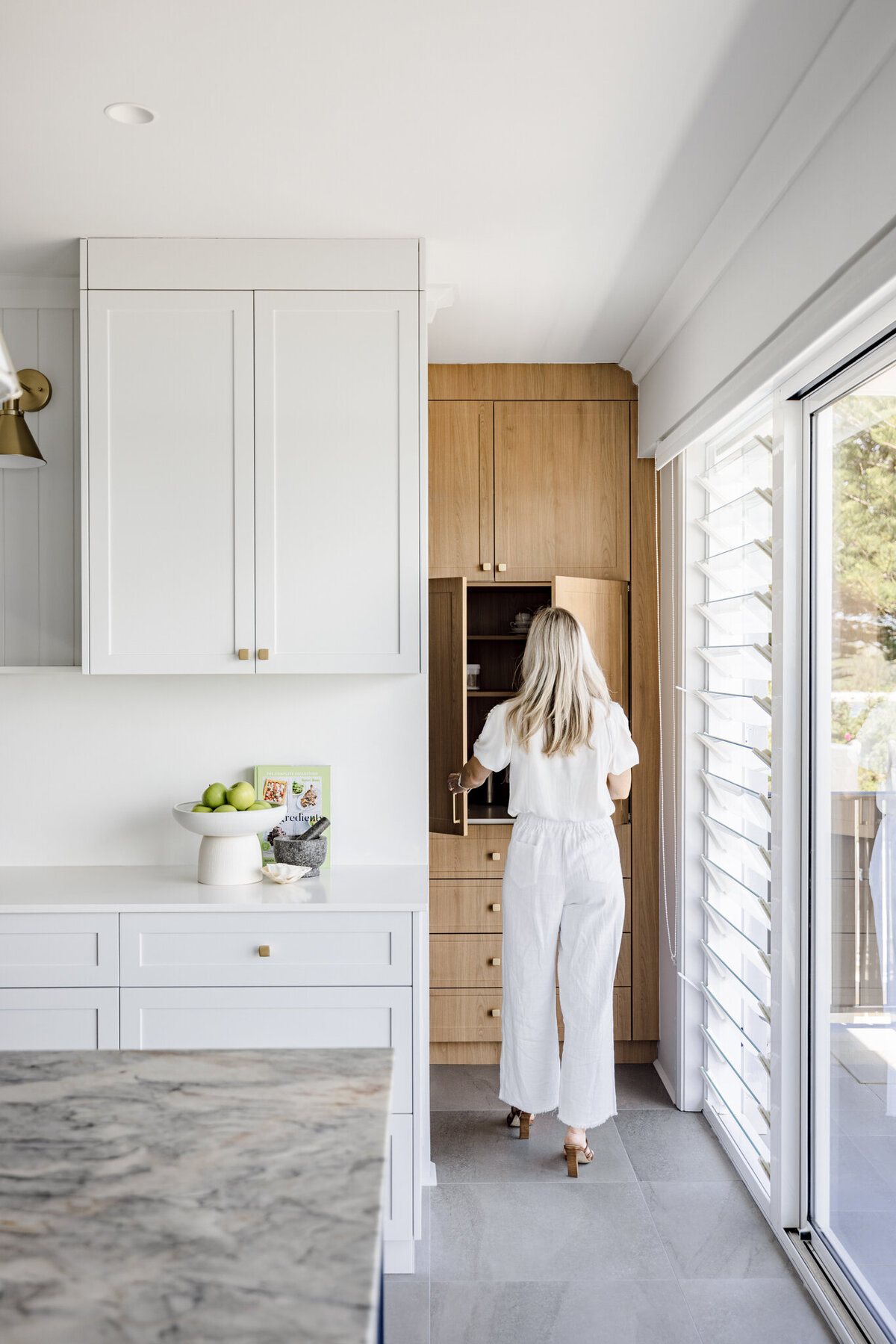 Person in white outfit opens wooden cabinet in a bright, luxury kitchen with a marble counter and large window, showcasing Newcastle Kitchen and Bathroom Designer's touch.