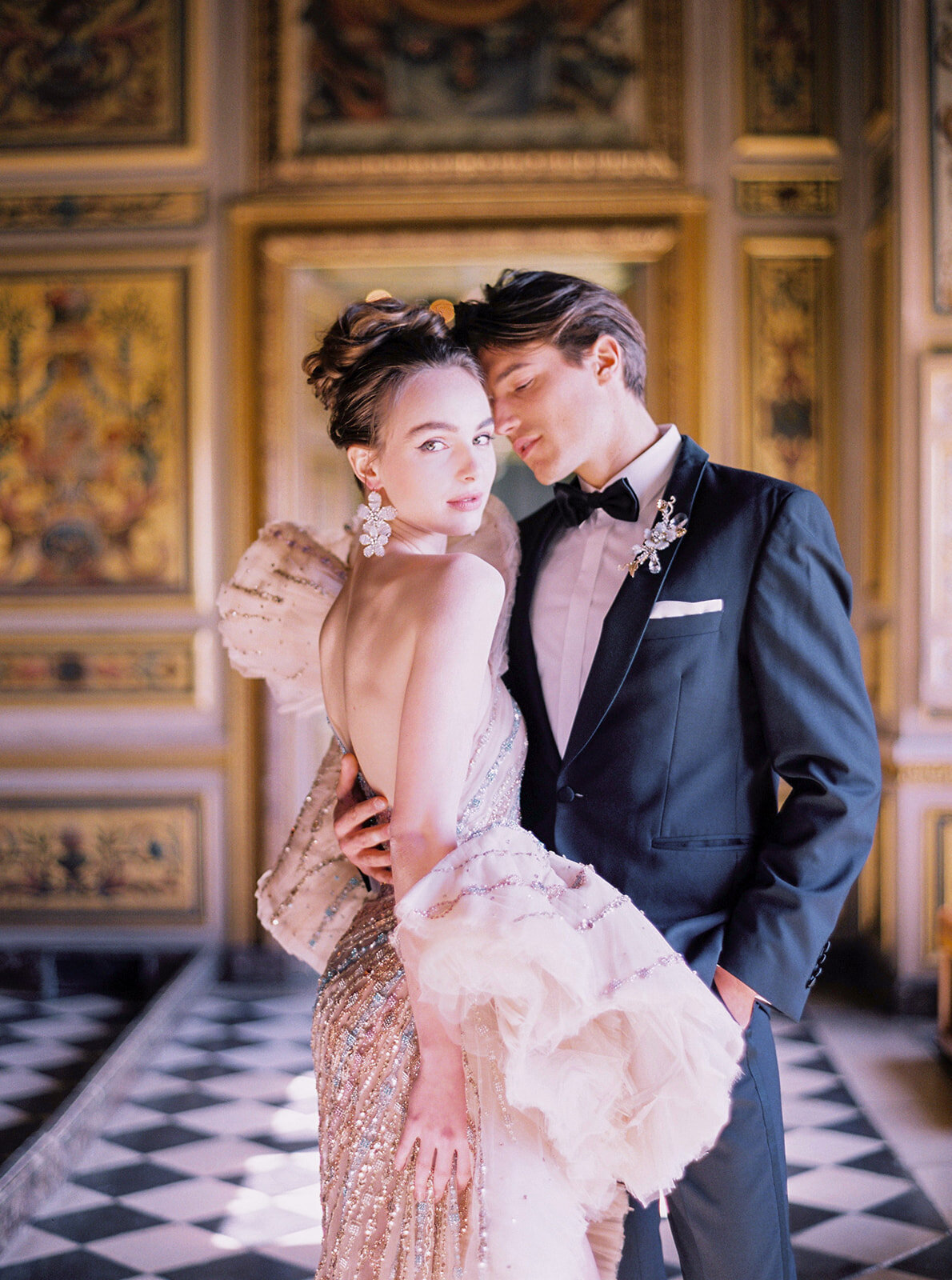 This stunning and elegant photo was captured at the stunning Chateau Vaux le Vicomte in Paris, France, making it a top destination wedding venue for couples looking for a romantic backdrop. The talented photographer Audrey perfectly captured the glamorous moment of a woman dressed in an embellished gown and large floral earrings standing closely with her partner in a classic black tuxedo with a bow tie. The ornate and opulent room with intricate golden details and checkered flooring adds a touch of sophistication to the overall ambiance, making it a truly magical setting for a special occasion.