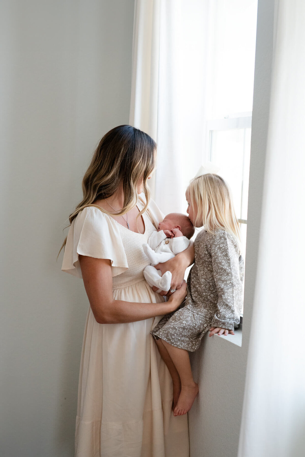 A toddler girl kisses her newborn sibling while sitting in a window sill with mom