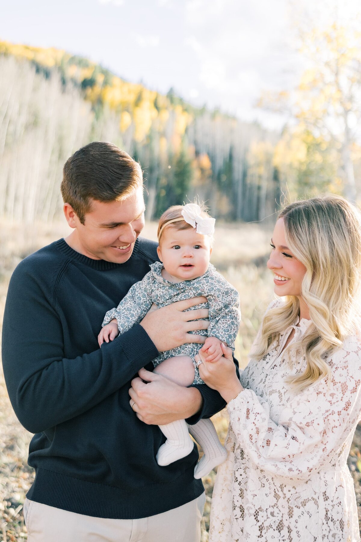 A young mom and dad are holding their 6 month old baby in their arms. There is a mountain scene with yellow aspens and mountains in the background. Taken by a local Breckenridge photographer. 