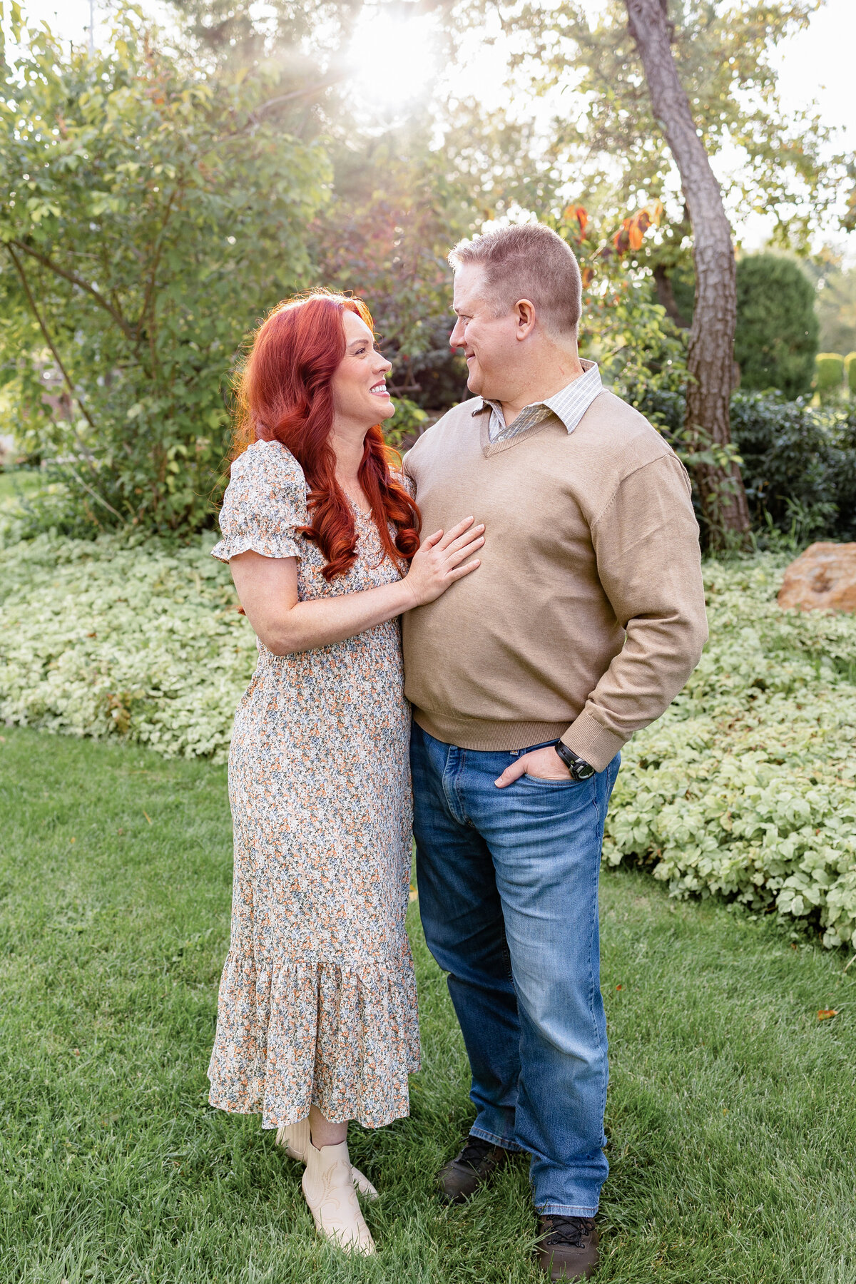 A couple stands close together, gazing lovingly at each other with soft sunlight filtering through the trees behind them.