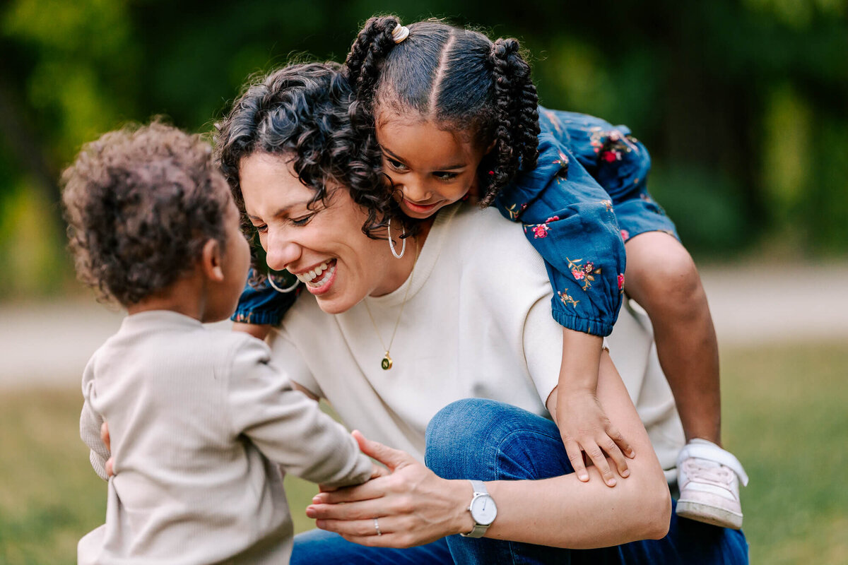 4 year old girl climbs on her mother's back, who is hunched on the ground taking her toddler's hands. Mom and little girl are laughing, their faces pressed close.