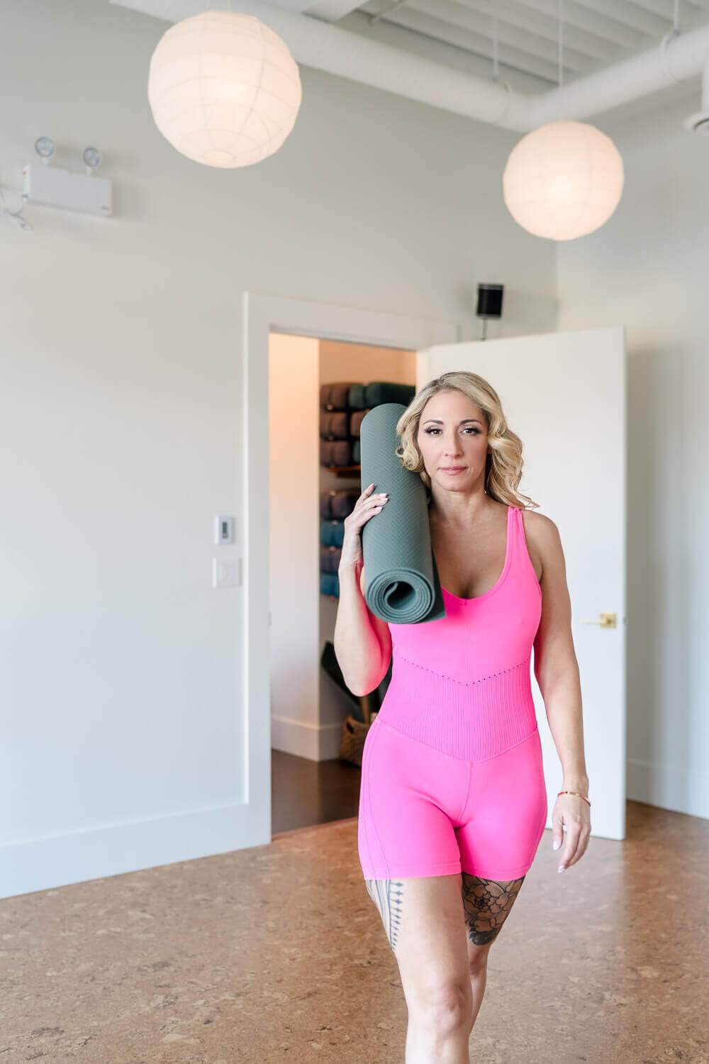 Professional woman walking towards camera carrying yoga mat over her shoulder in bright modern yoga studio.