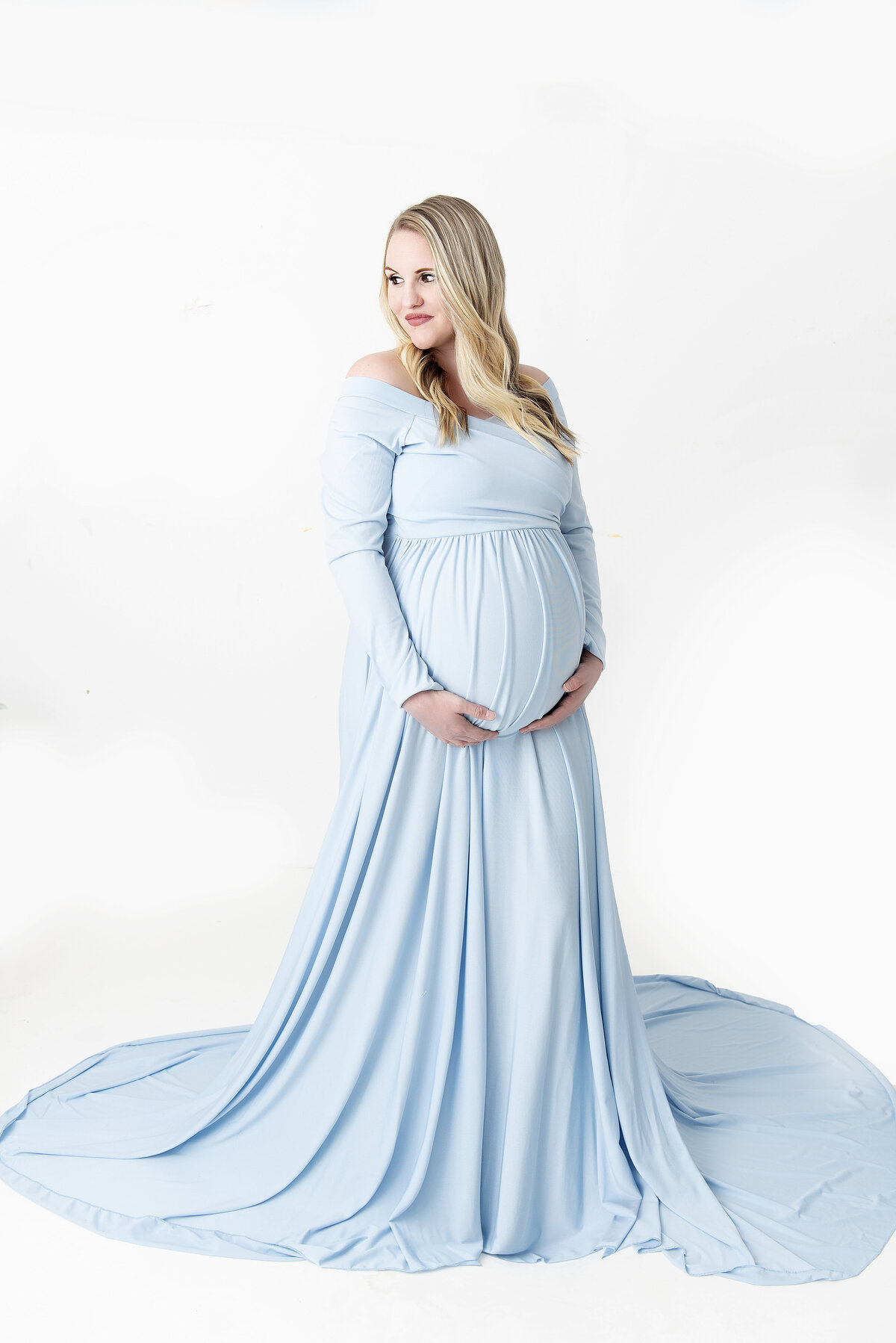 A blonde mother to be holds er bump while standing in a studio looking over her shoulder in a long blue maternity gown