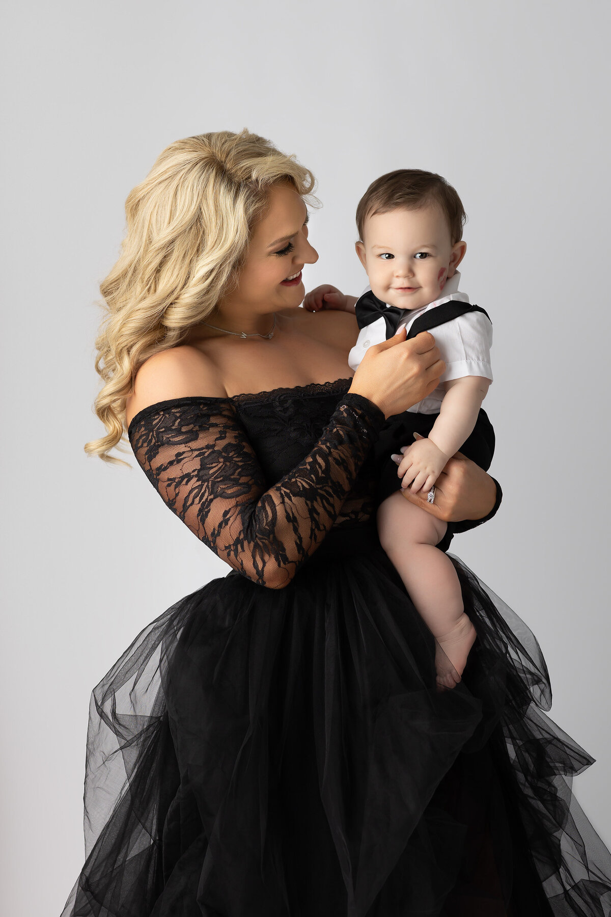 A baby in a tuxedo onesie with a lipstick kiss on his cheek looks confidently at the camera while being held by his smiling mom in a black lace gown, captured by a Dallas milestone photographer.