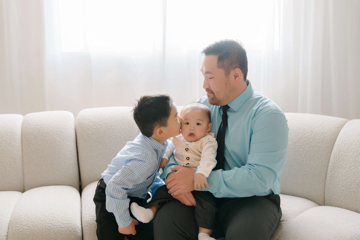 Big brother leans in to kiss his baby brother while their father looks on lovingly in a light-filled studio.