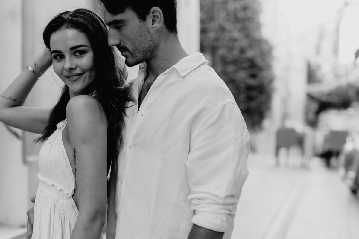 Couple standing together with her back against him as she looks at the camera and he looks at her.