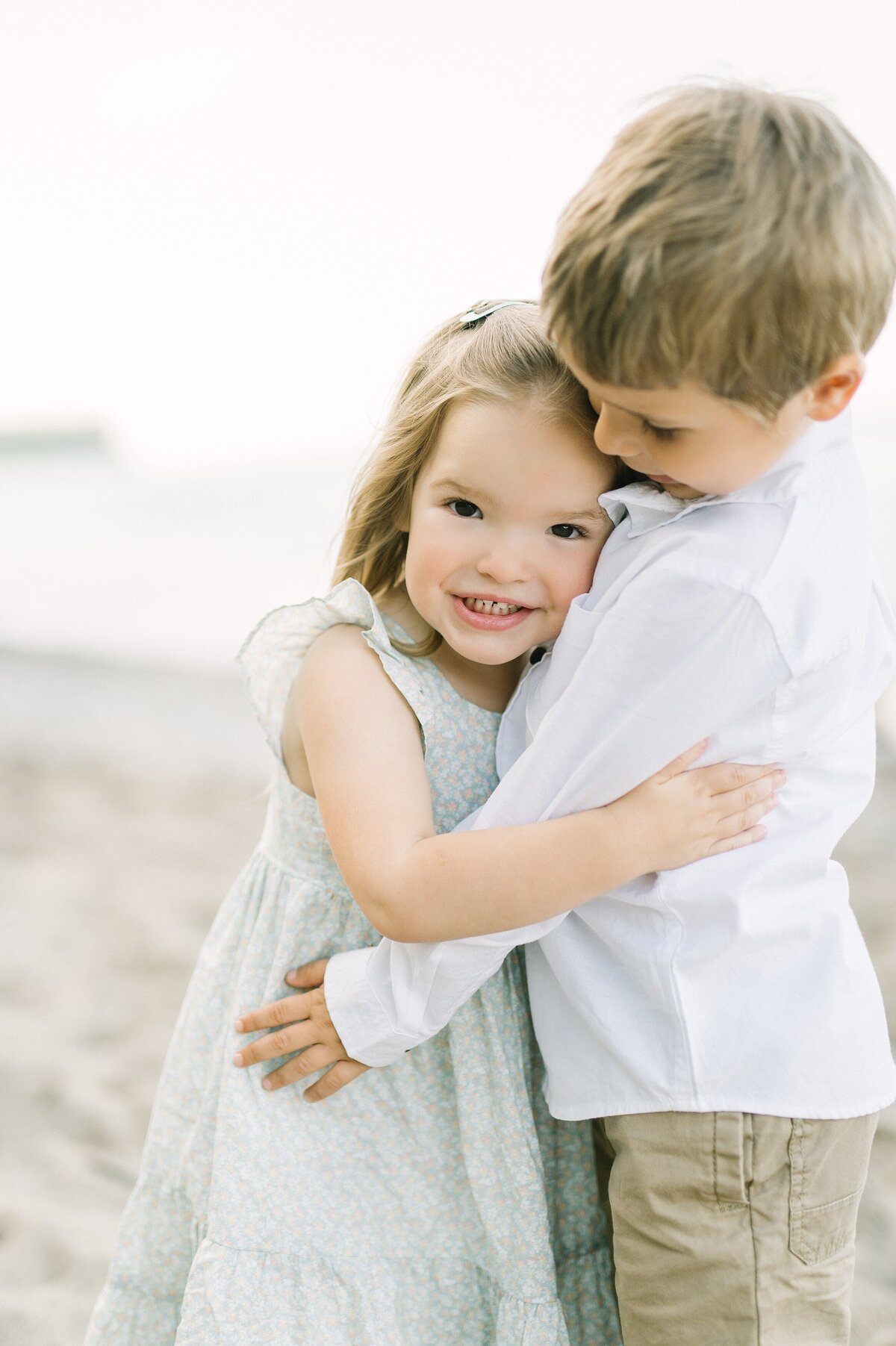 Two siblings hugging on the beach, with the brother smiling at his sister
