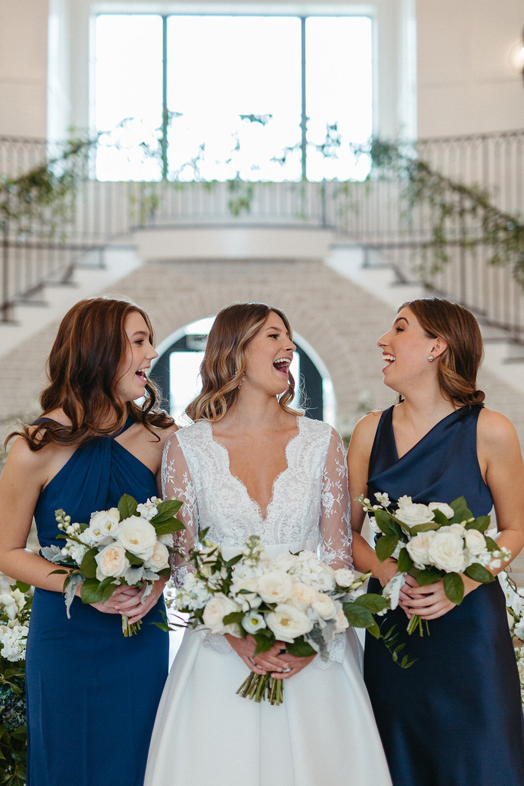 A bride in a white wedding gown holding a white bouquet smiles between two bridesmaids wearing blue satin gowns.