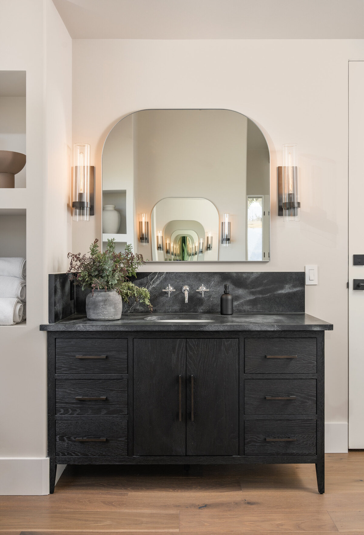 black vanity soapstone countertop arched mirror restoration hardware sconces gray vase with branches styled built-ins wall mount faucet\