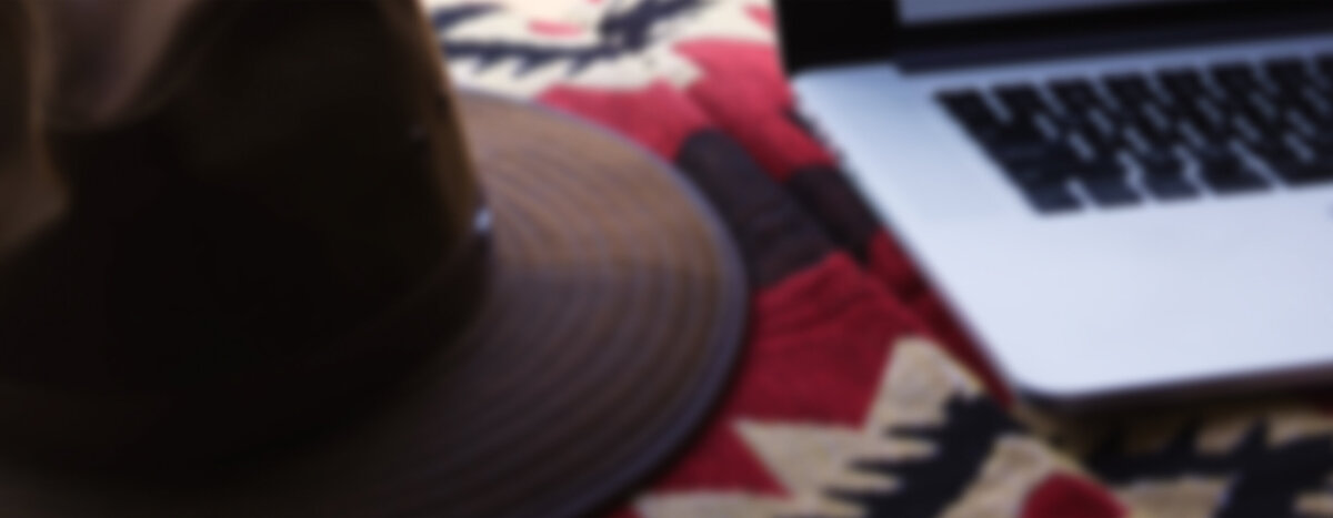 Blurry brown hat and grey laptop sitting on a red tribal blanket.