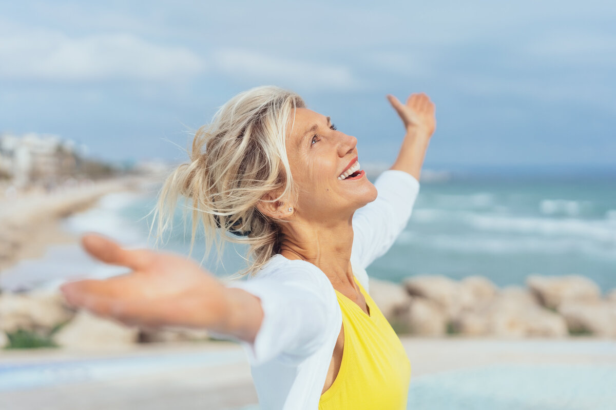 Older, smiling woman in yellow shirt looks to the sky with arms open.