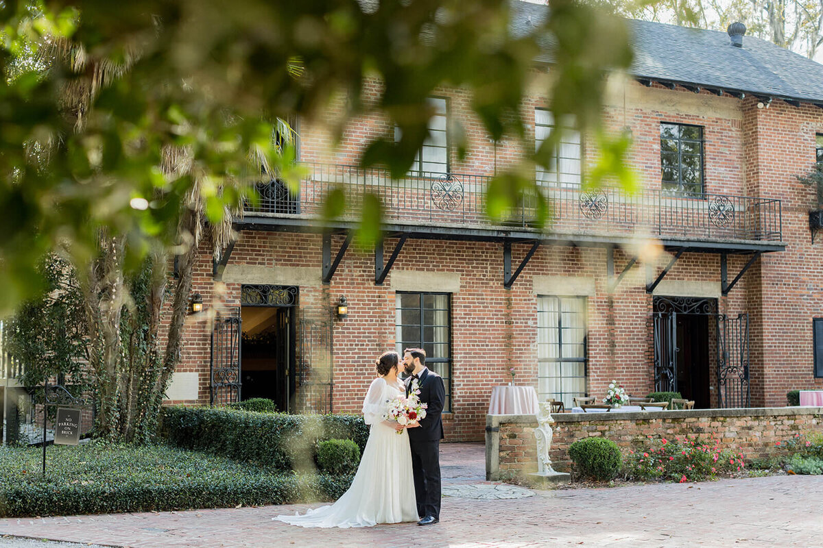 A romantic moment at Millstone at Adams Pond, where a newlywed couple shares a quiet embrace against the backdrop of a historic brick estate. Lush greenery frames the scene, adding depth and intimacy to this dreamy Southern wedding portrait. Captured by South Carolina luxury wedding photographer Jessica Hunt Photography.