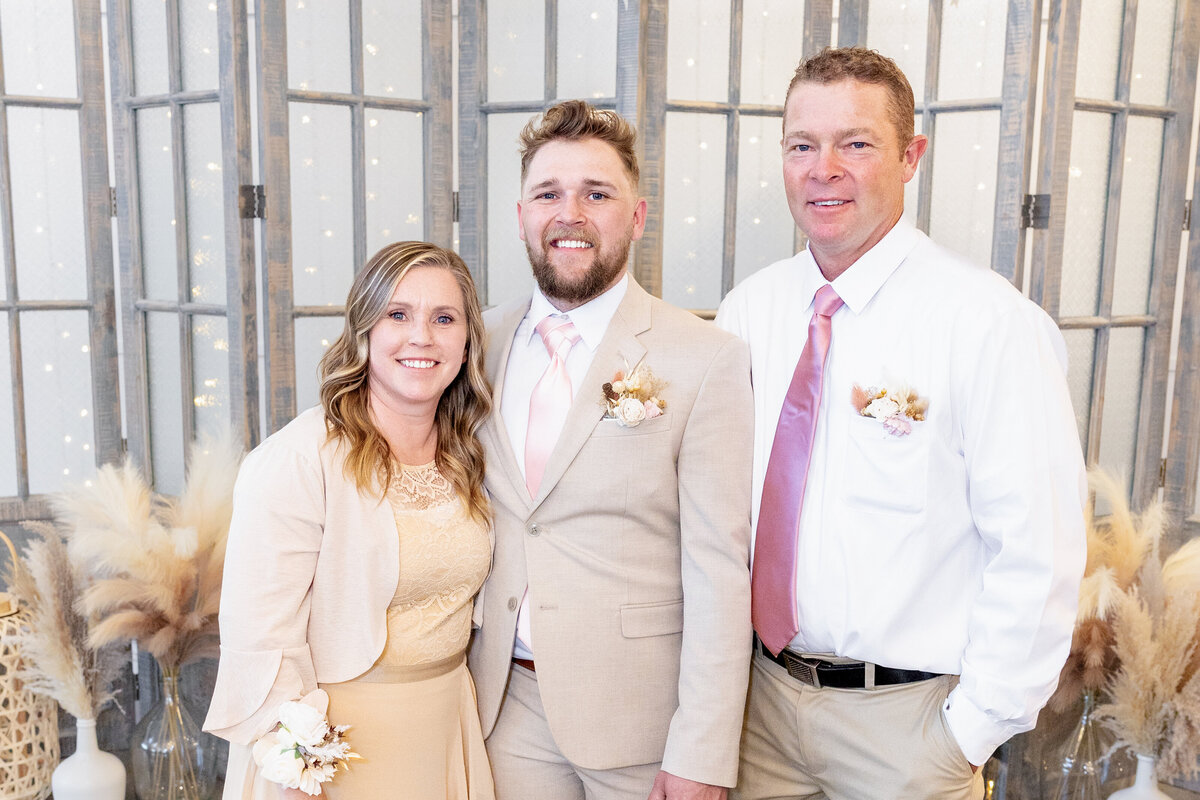 The groom poses proudly with his parents, celebrating the special day with a heartfelt family moment. Dressed in neutral, elegant tones, the family stands in front of the beautifully decorated altar at Maplewood Events in Spanish Fork, Utah, captured by Cassie Lee Photography.