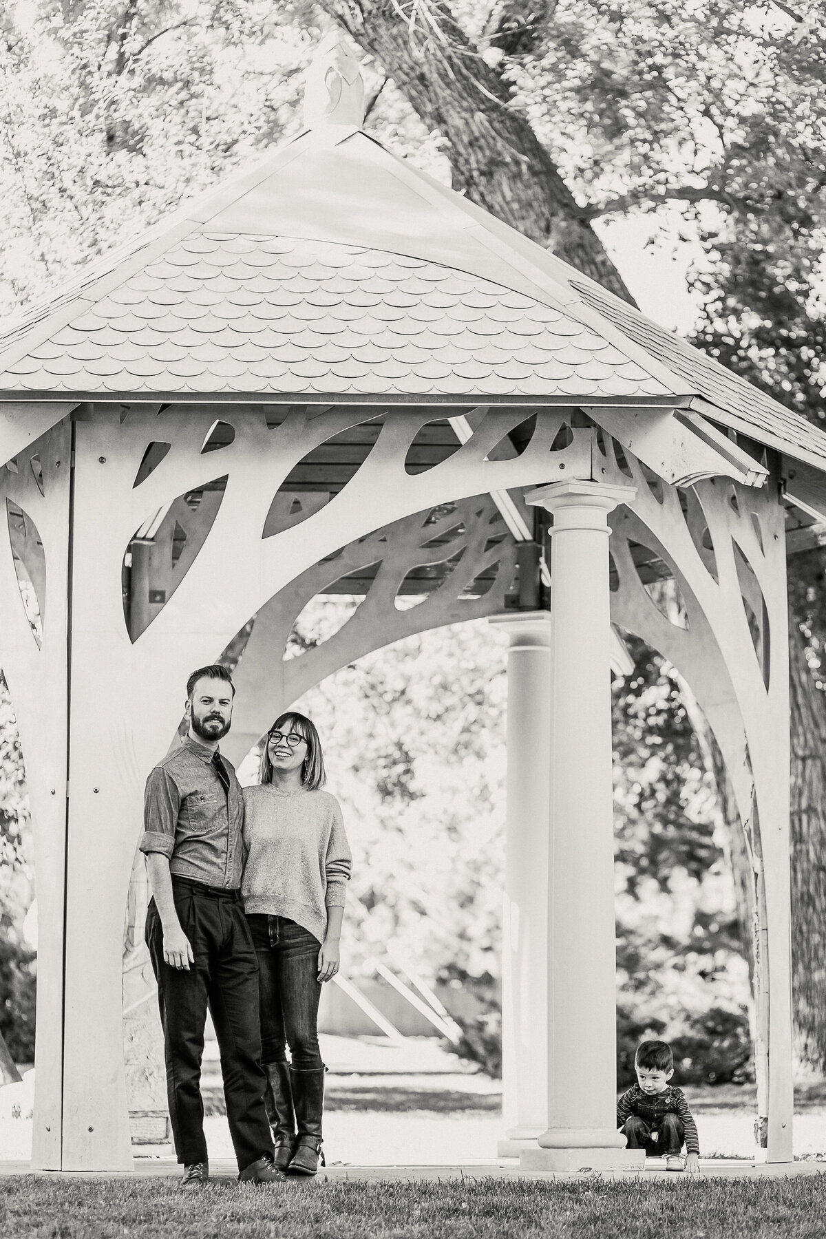 black and white photo of parents standing in front of gazebo while son crouches behind pole