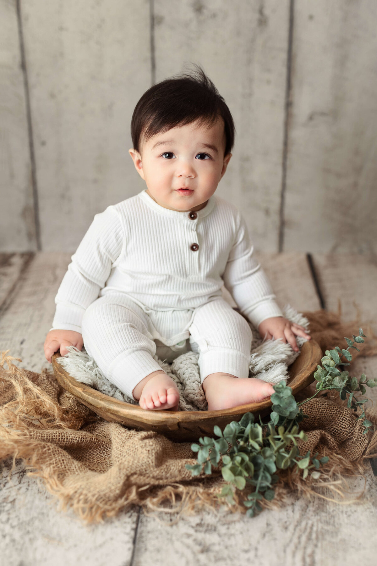 6 moth old boy in a white outfit sitting on a wooden bowl with greenery around it on a white wooden backdrop during a baby milestone session