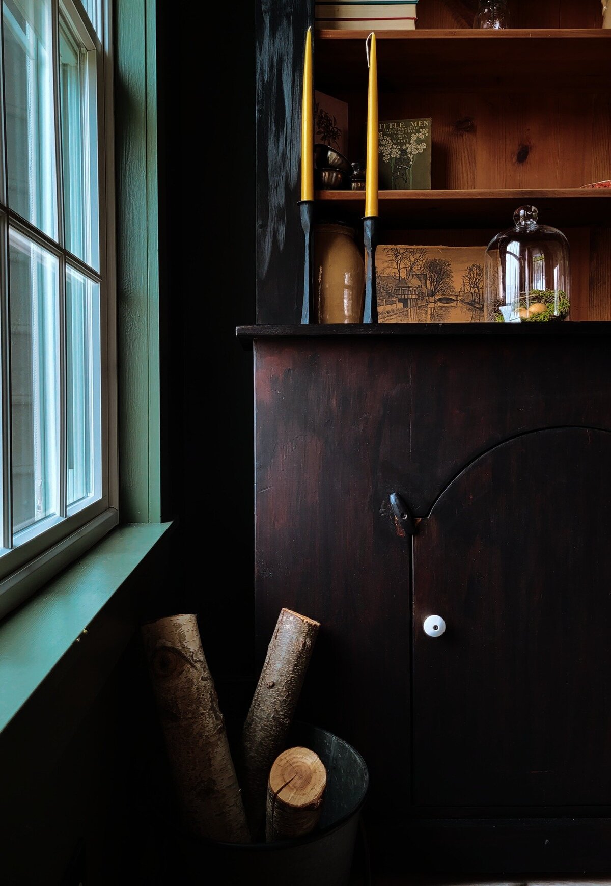 Black library cabinet with arched door cabinet base door with tin bucket containing birch logs, beside a window