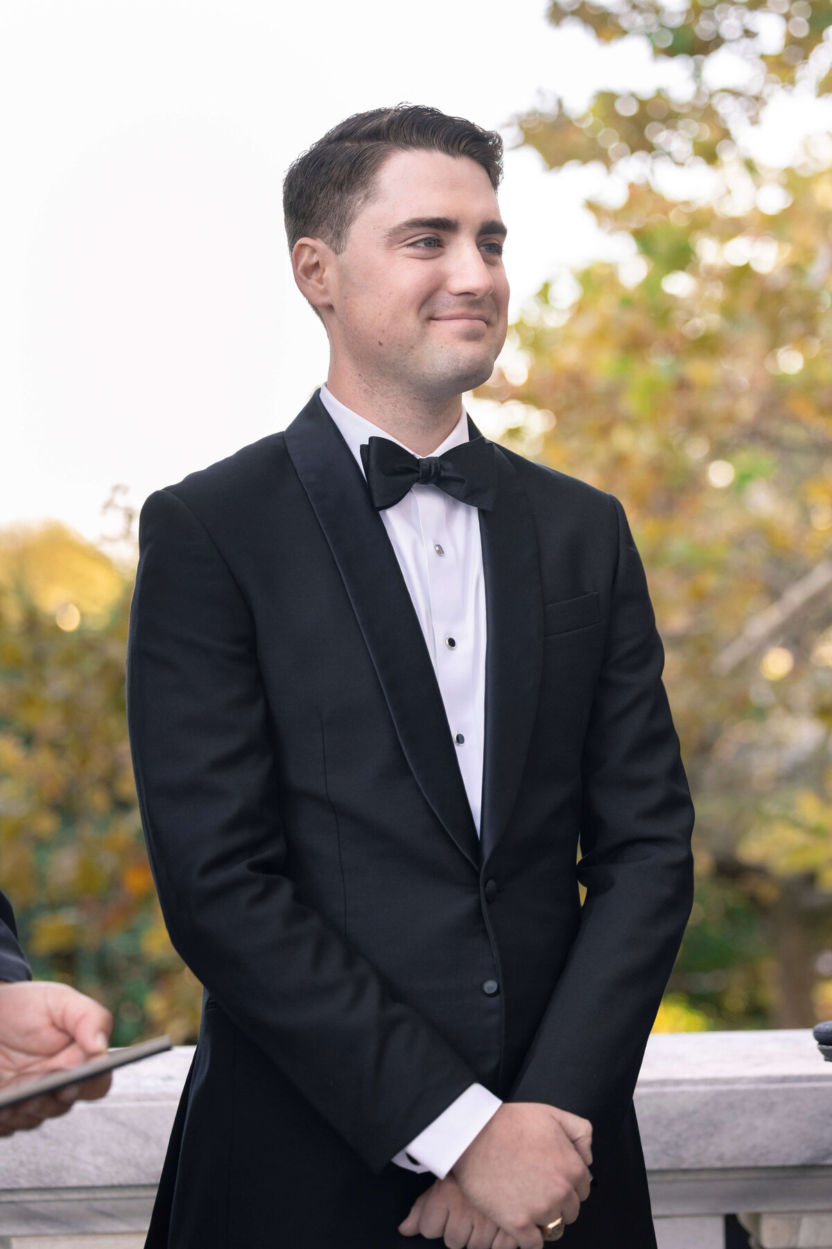 A man in a black tuxedo and bow tie stands outdoors, smiling softly. The background features blurred autumn leaves and a white railing, suggesting a formal event or ceremony.