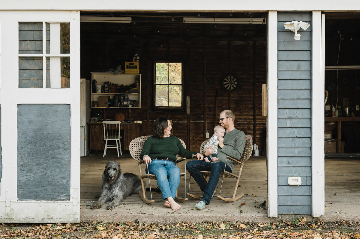 Couple sitting on chairs in front of barn with dogs and baby.