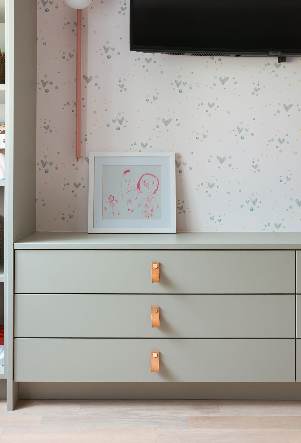 A pastel green dresser in a cozy children's playroom. The dresser has brown leather pulls. On top of the dresser is a framed piece of children's artwork.