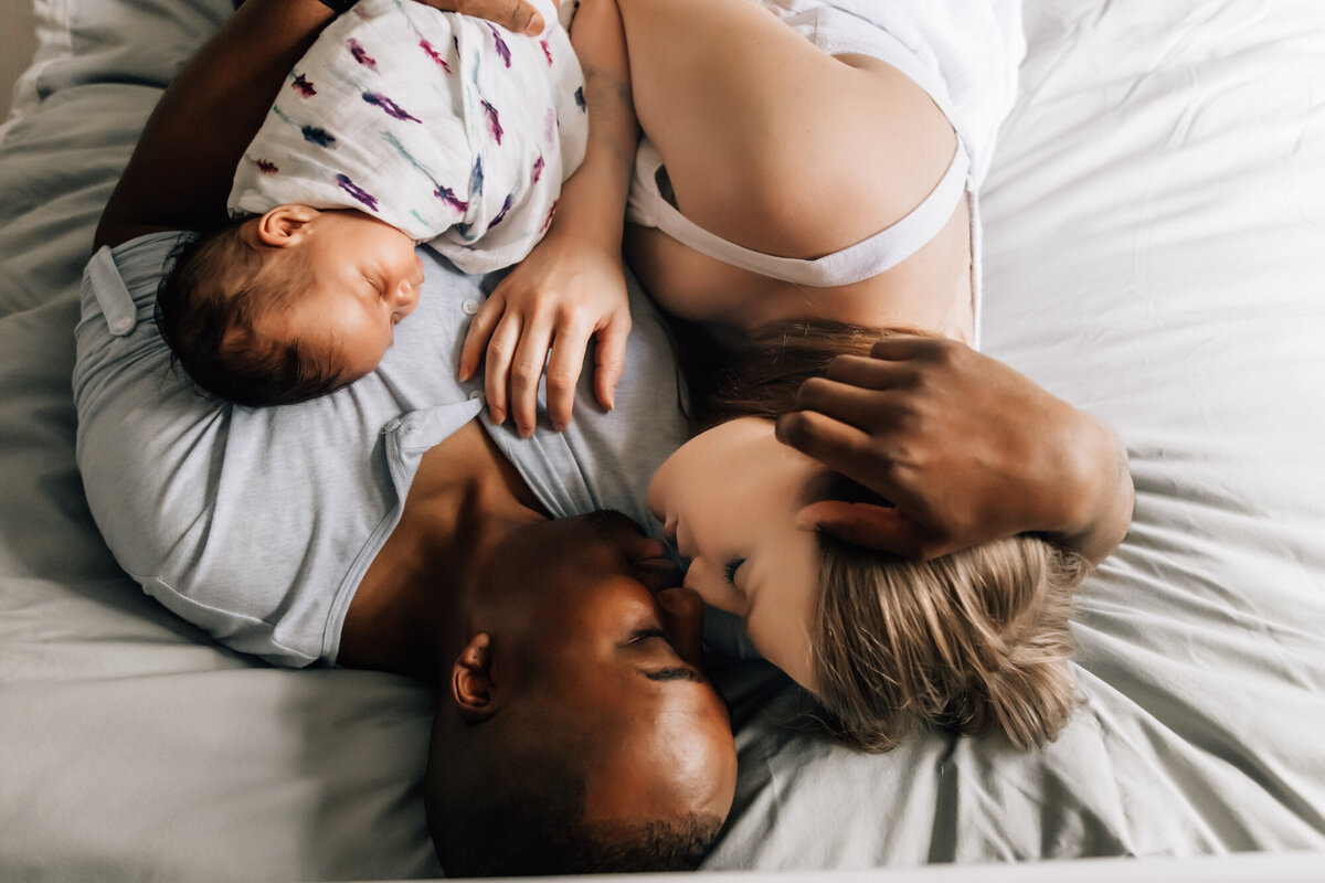 Newborn Photographer, Mom and dad laying on the bed kissing while their baby girl is asleep on dad's chest.