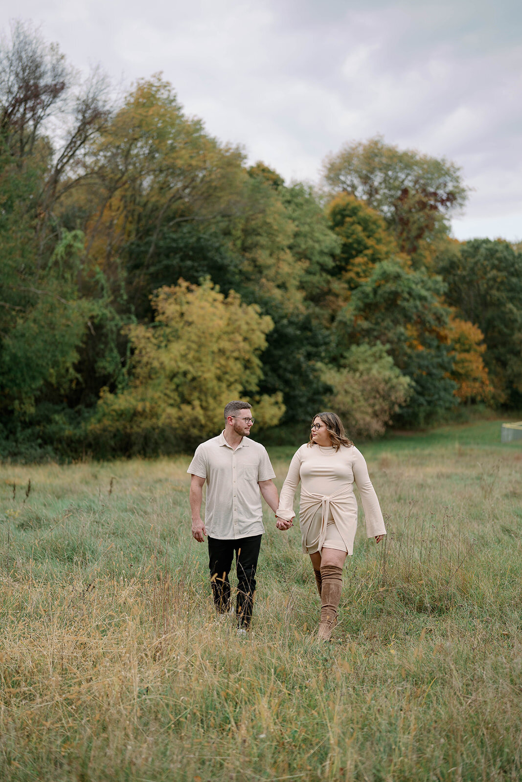 Engaged couple walking together through an open grassy field surrounded by fall colors at Al Sabo Preserve.