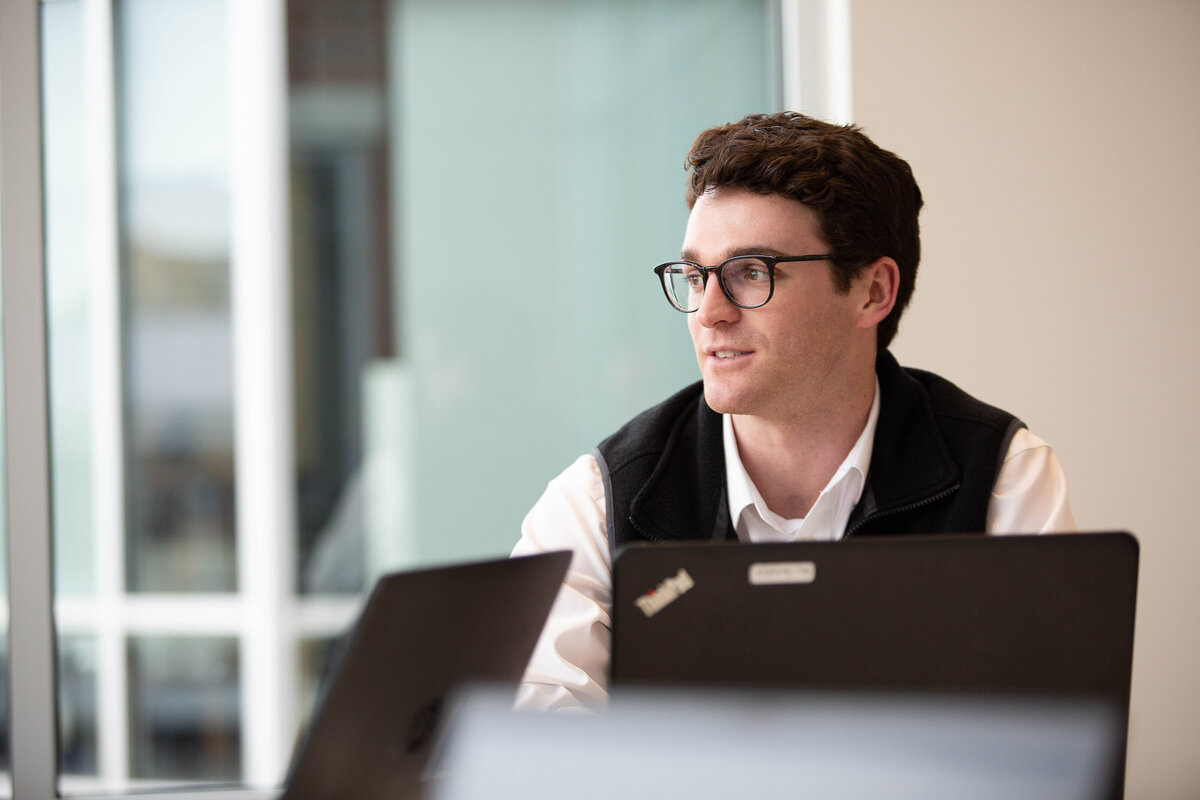 Young white male wearing glasses looking to the left with an open laptop in front of him.