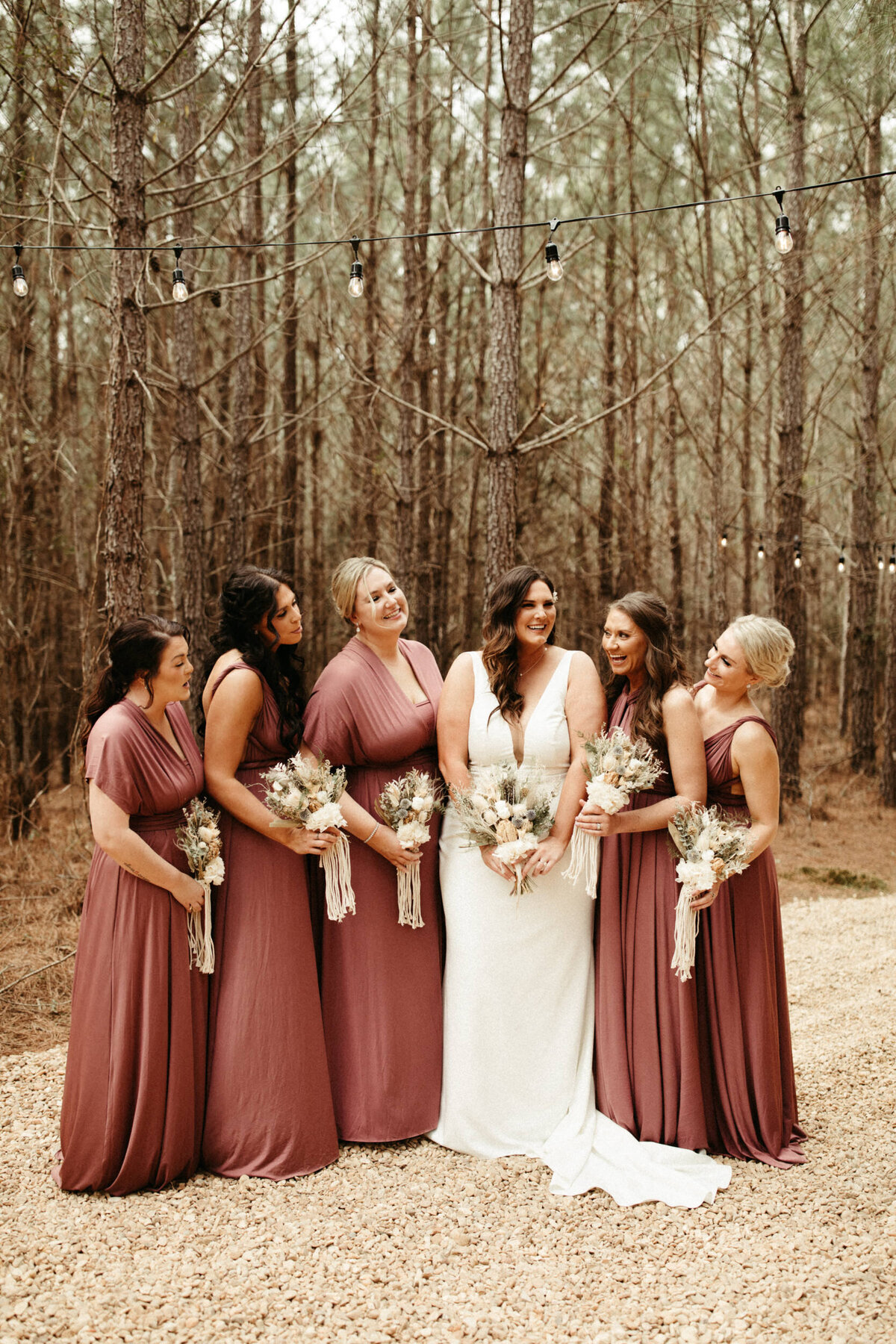 Boho bride with her bridesmaids wearing burgundy dresses and holding dried floral bouquets standing in the woods on a winter day with string lights hanging over them