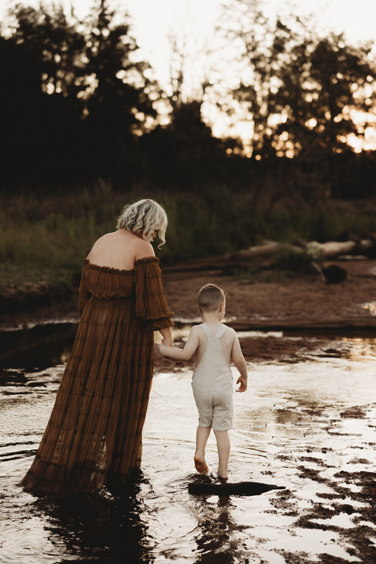 A Mum and a child wade through a shallow body of water with a forested background at sunset. The adult wears a long brown dress while the child is in a light-colored outfit, capturing the serene essence of South Coast family photography.