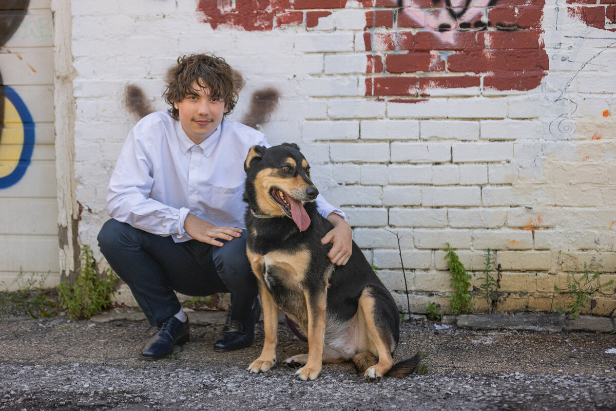A senior guy squatting next to his dog against a brick wall in Lawrence, KS