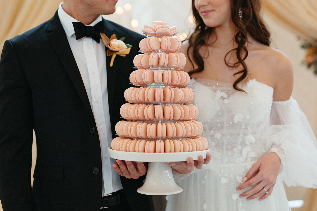 Close-up of bride and groom wearing a black tuxedo and white wedding gown holding a cake stand filled with macaroons.